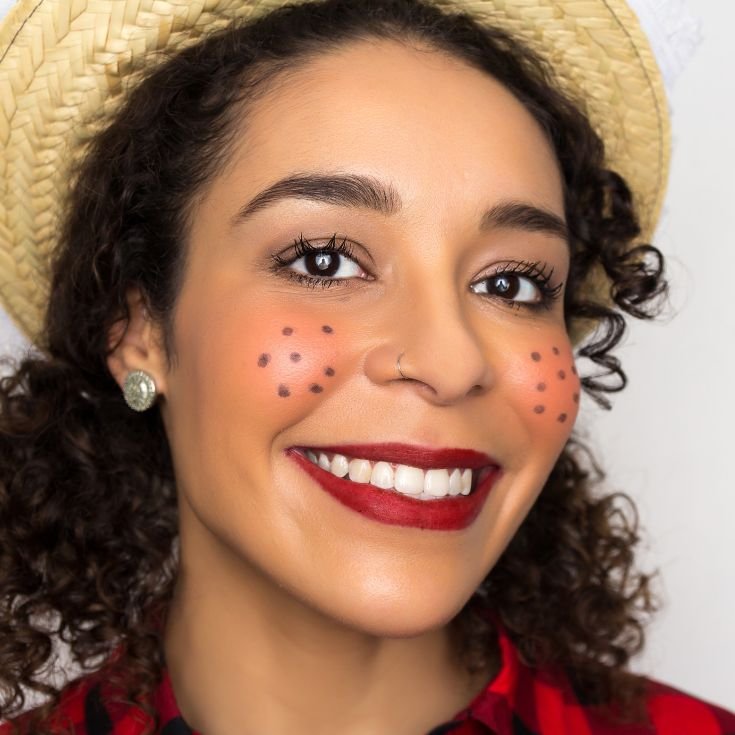 ulher de cabelo cacheado utilizando chapéu de palha e usando maquiagem tipicamente junina, com blush intenso e sardinhas de lápis de olho preto.