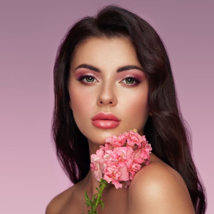Mulher usando maquiagem leve e rosada, com tons mais claros no meio das pálpebras, sendo esfumado para cores mais escuras nas extremidades. A pele está impecável e em sua boca está um batom rosa médio com acabamento brilhante.