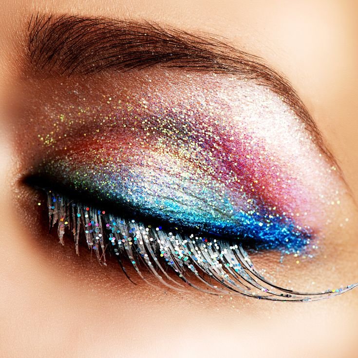 Olho maquiado com sombras coloridas, com azul na linha dos cílios, e amarelo e rosa no exterior. Todo o olho tem glitter, e a pessoa usa cílios postiços longos