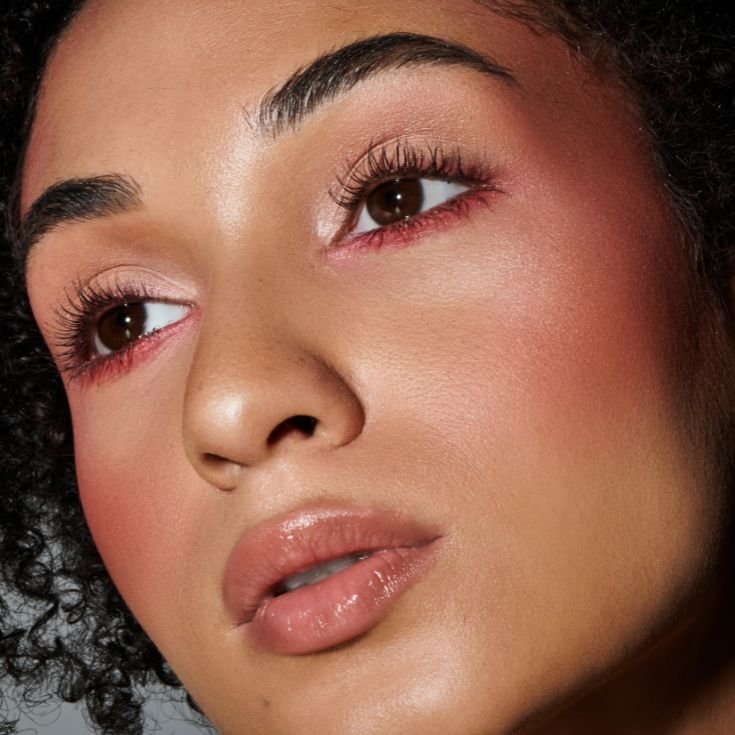 Mulher jovem com maquiagem rosada nos olhos, blush suave, lábios glossy e cabelo cacheado. Olhar direcionado para cima. Pele iluminada.