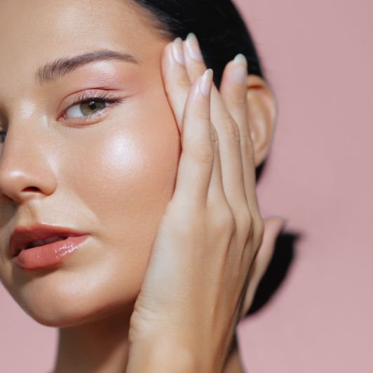 Mulher com pele iluminada e maquiagem natural em tons rosados, lábios glossy, cabelo escuro preso, tocando o rosto delicadamente com a mão em um fundo rosa.