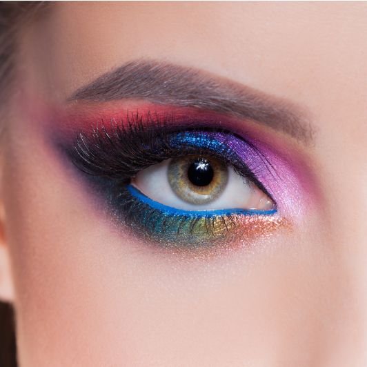 Mulher de olhos verdes usando sombras cintilantes  nas cores azul, roxo, rosa, dourado e verde com um lápis de olho azul na linha d’água e sobrancelha marcada.