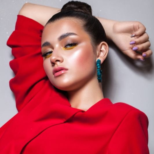Mulher com maquiagem marcante com sombra amarela, pele iluminada, blush rosado, brinco turquesa, unhas lilás e cabelo preso em coque posando em um fundo cinza.