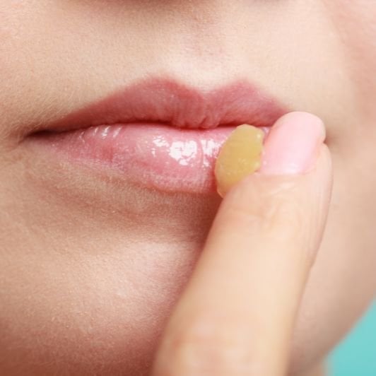Close de lábios naturais com brilho glossy enquanto o dedo aplica um bálsamo labial hidratante, destacando a textura macia e saudável da pele.