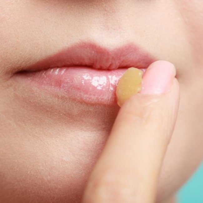 Close de lábios naturais com brilho glossy enquanto o dedo aplica um bálsamo labial hidratante, destacando a textura macia e saudável da pele.